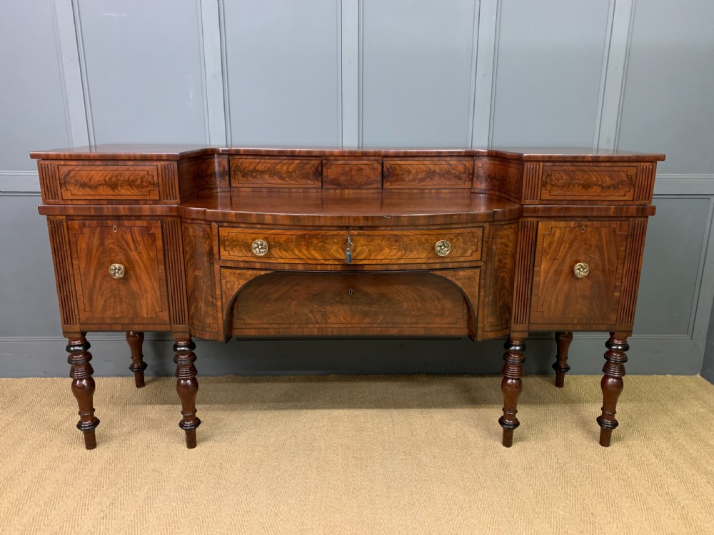 A Regency period flame mahogany sideboard featuring a raised stage top, central drawer, two side cupboards, detailed stringing, and turned reeded legs. View II