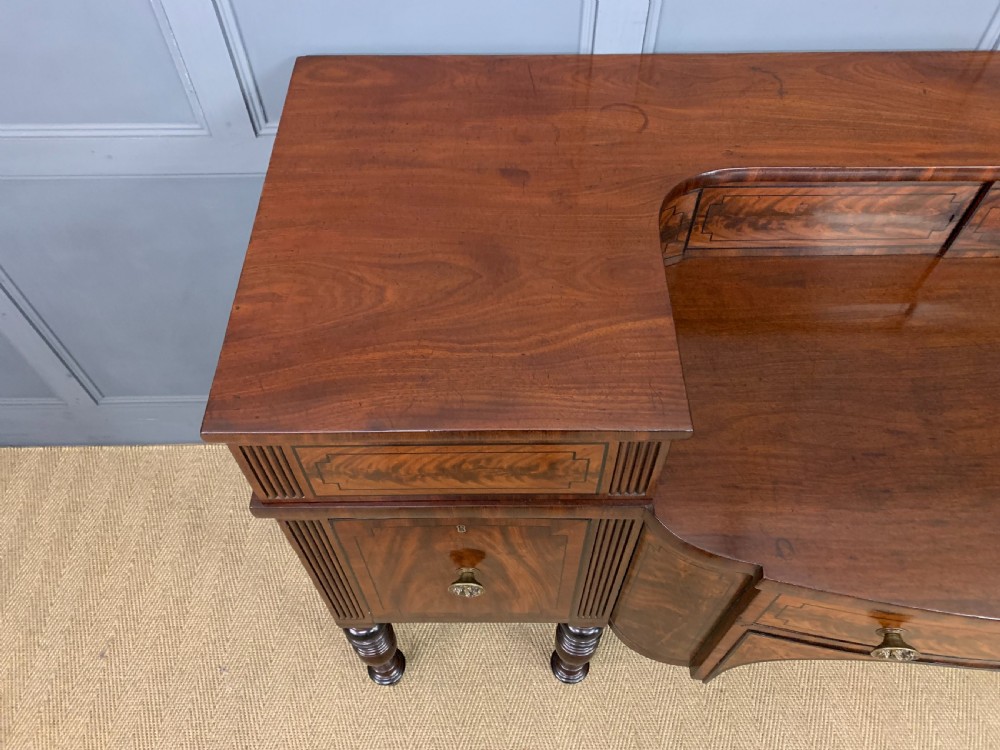 A Regency period flame mahogany sideboard featuring a raised stage top, central drawer, two side cupboards, detailed stringing, and turned reeded legs. View III