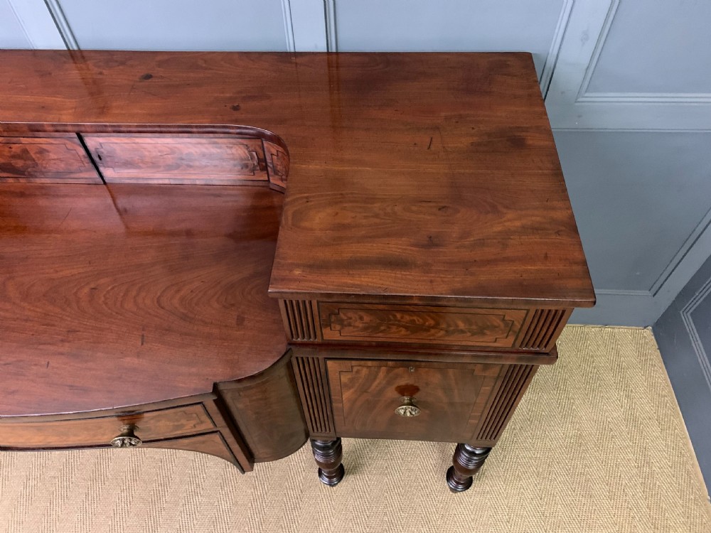 A Regency period flame mahogany sideboard featuring a raised stage top, central drawer, two side cupboards, detailed stringing, and turned reeded legs. View IV