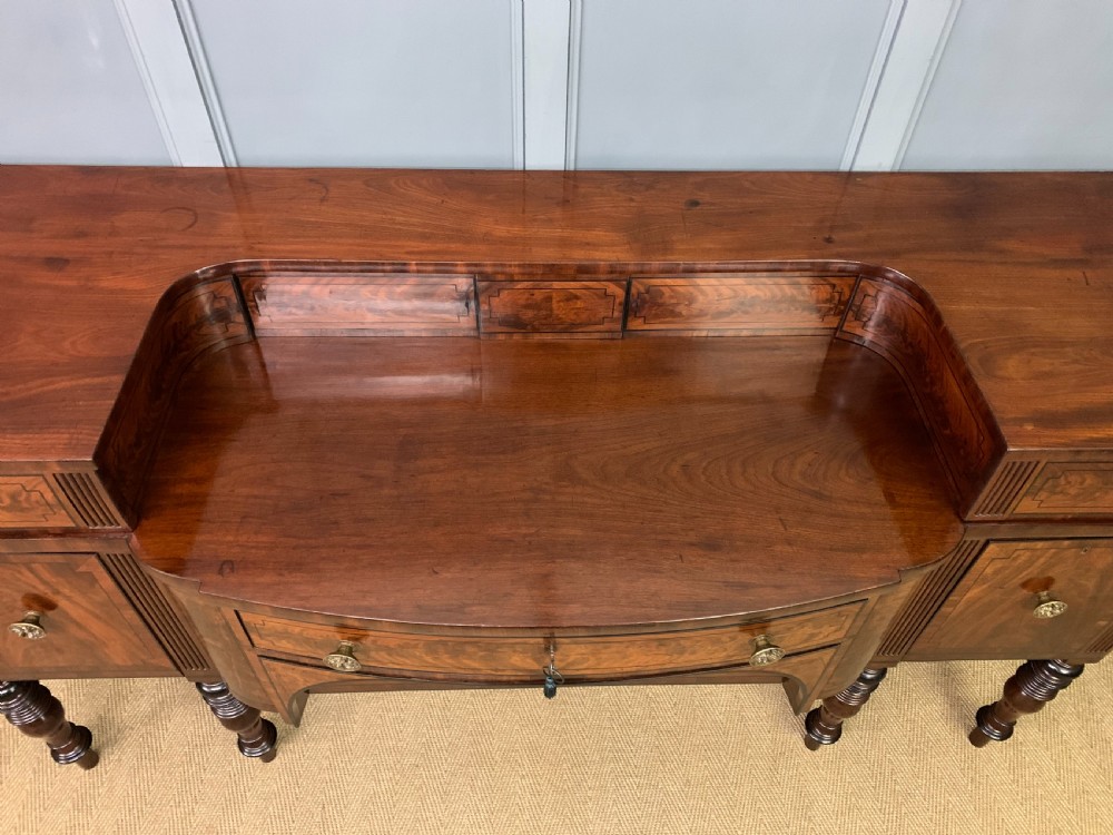 A Regency period flame mahogany sideboard featuring a raised stage top, central drawer, two side cupboards, detailed stringing, and turned reeded legs. View V
