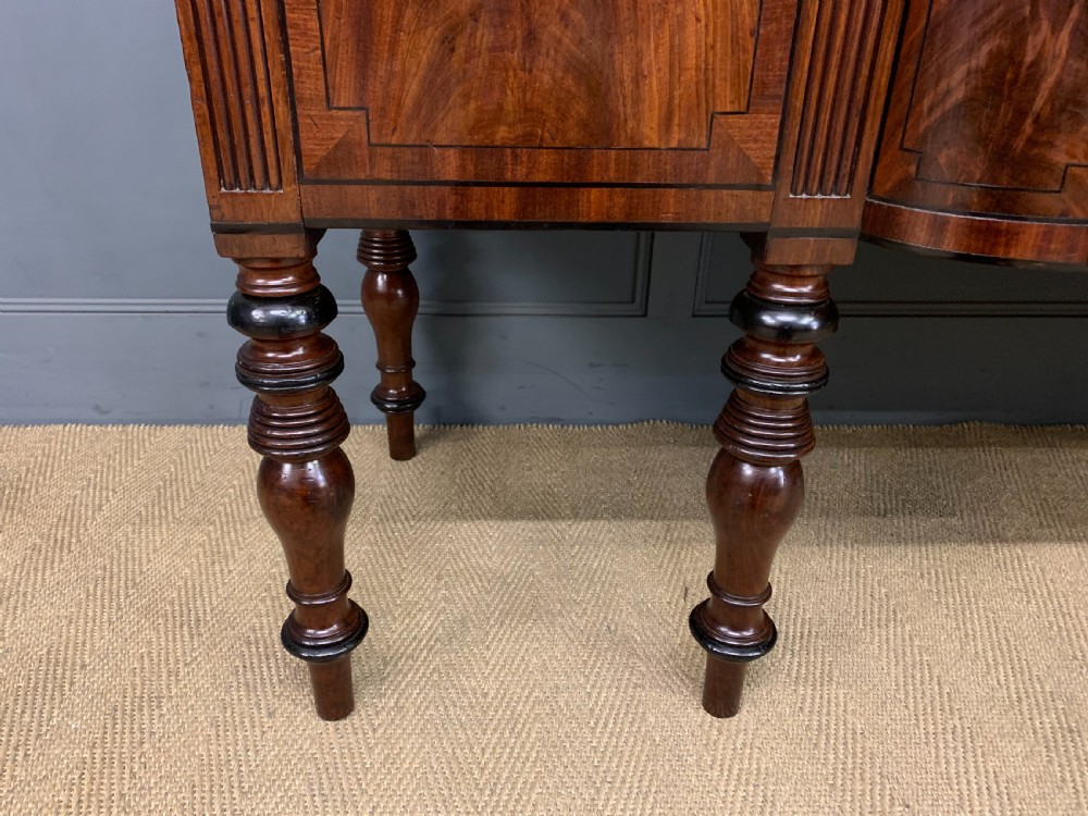 A Regency period flame mahogany sideboard featuring a raised stage top, central drawer, two side cupboards, detailed stringing, and turned reeded legs. View VI