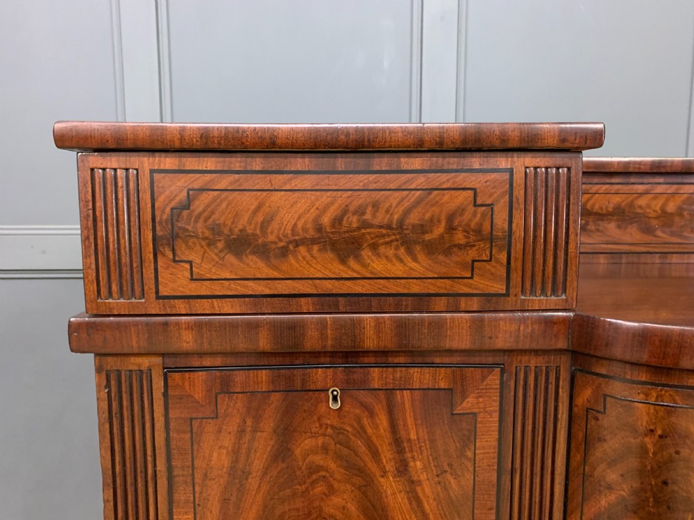 A Regency period flame mahogany sideboard featuring a raised stage top, central drawer, two side cupboards, detailed stringing, and turned reeded legs. View VII