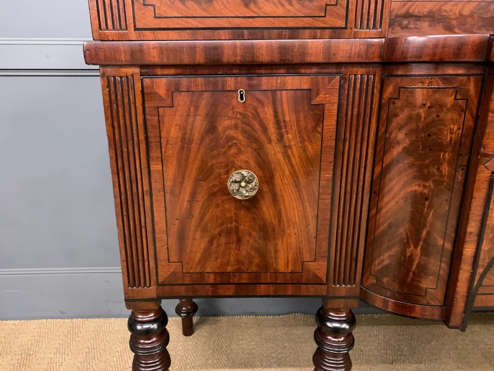 A Regency period flame mahogany sideboard featuring a raised stage top, central drawer, two side cupboards, detailed stringing, and turned reeded legs. View VIII