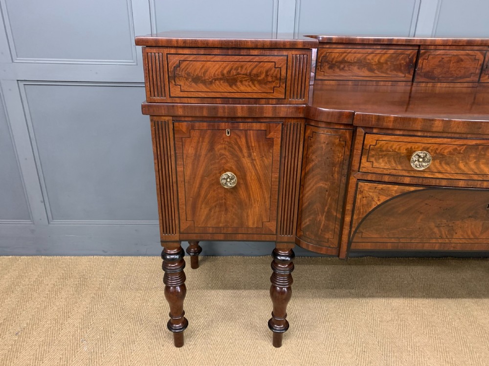 A Regency period flame mahogany sideboard featuring a raised stage top, central drawer, two side cupboards, detailed stringing, and turned reeded legs. View X