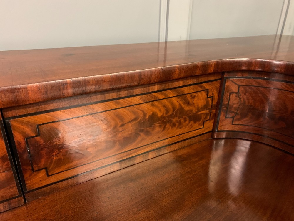 A Regency period flame mahogany sideboard featuring a raised stage top, central drawer, two side cupboards, detailed stringing, and turned reeded legs. View XII