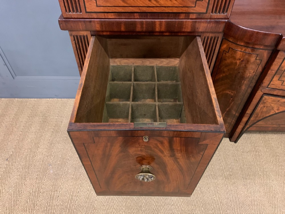 A Regency period flame mahogany sideboard featuring a raised stage top, central drawer, two side cupboards, detailed stringing, and turned reeded legs. View XIV