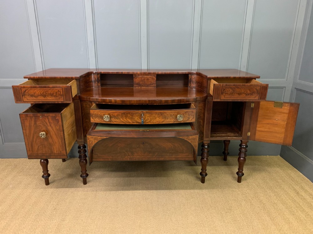 A Regency period flame mahogany sideboard featuring a raised stage top, central drawer, two side cupboards, detailed stringing, and turned reeded legs. View XV