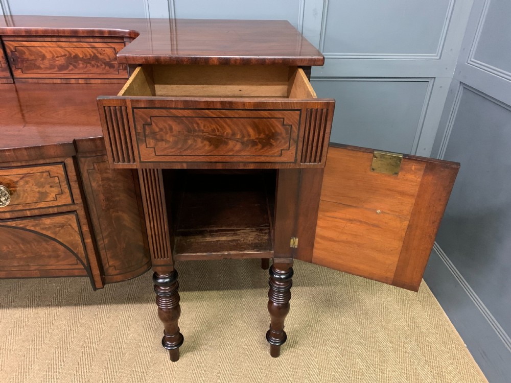 A Regency period flame mahogany sideboard featuring a raised stage top, central drawer, two side cupboards, detailed stringing, and turned reeded legs. View XVII