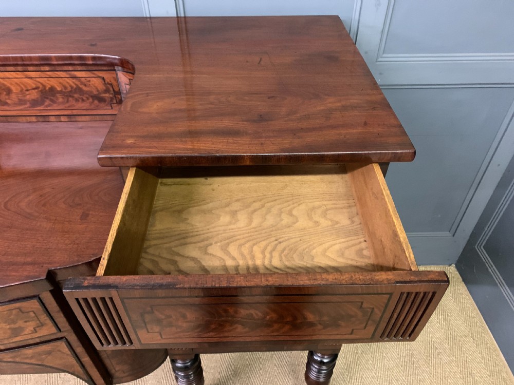 A Regency period flame mahogany sideboard featuring a raised stage top, central drawer, two side cupboards, detailed stringing, and turned reeded legs. View XVIII