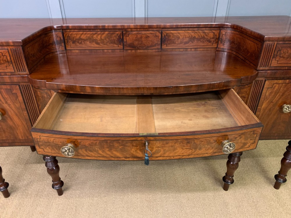 A Regency period flame mahogany sideboard featuring a raised stage top, central drawer, two side cupboards, detailed stringing, and turned reeded legs. View XX