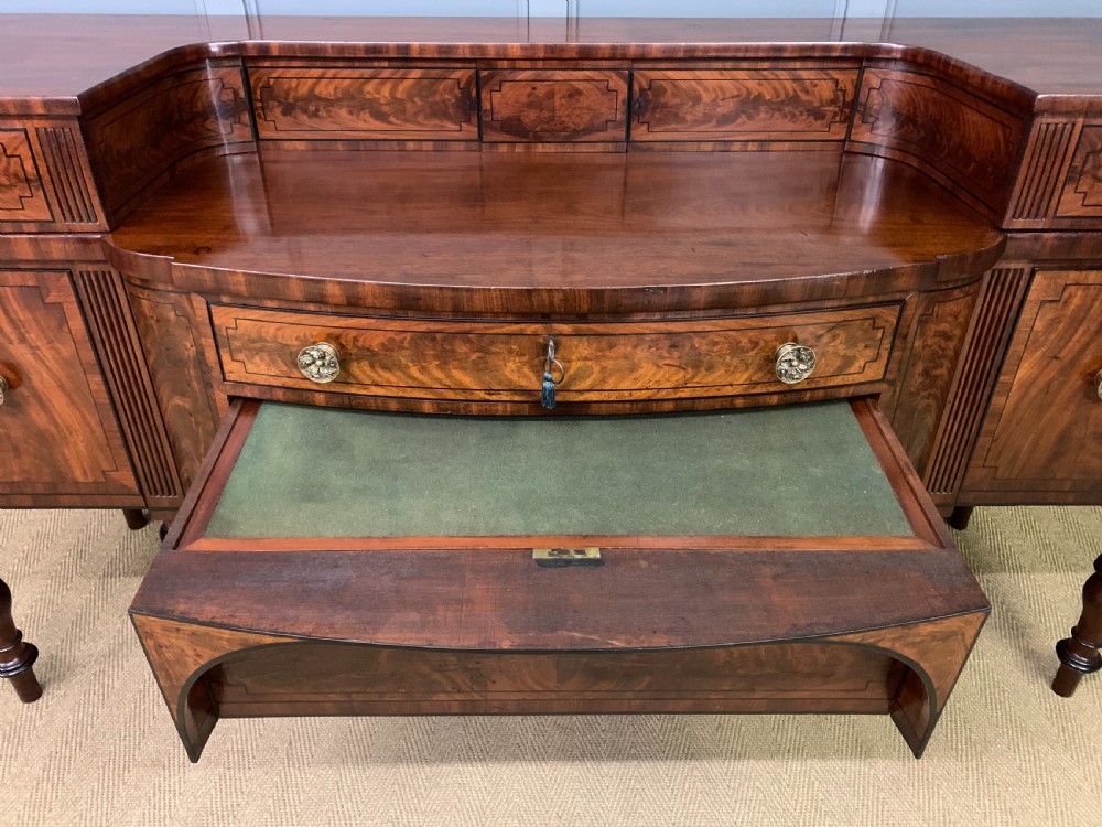 A Regency period flame mahogany sideboard featuring a raised stage top, central drawer, two side cupboards, detailed stringing, and turned reeded legs. View XXI