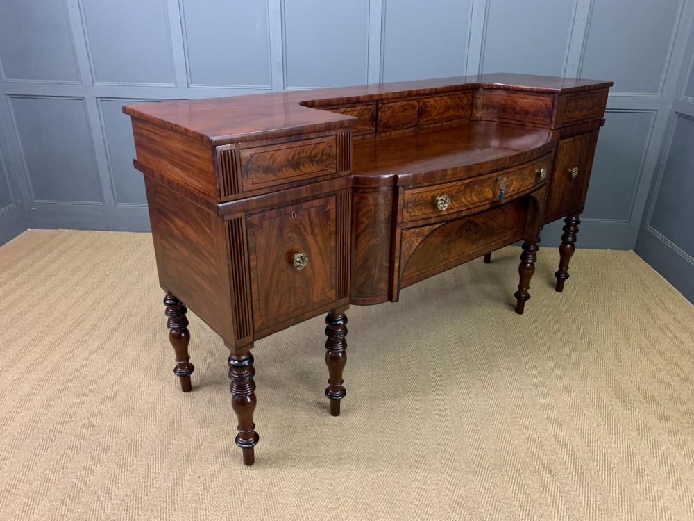A Regency period flame mahogany sideboard featuring a raised stage top, central drawer, two side cupboards, detailed stringing, and turned reeded legs. View XXIII