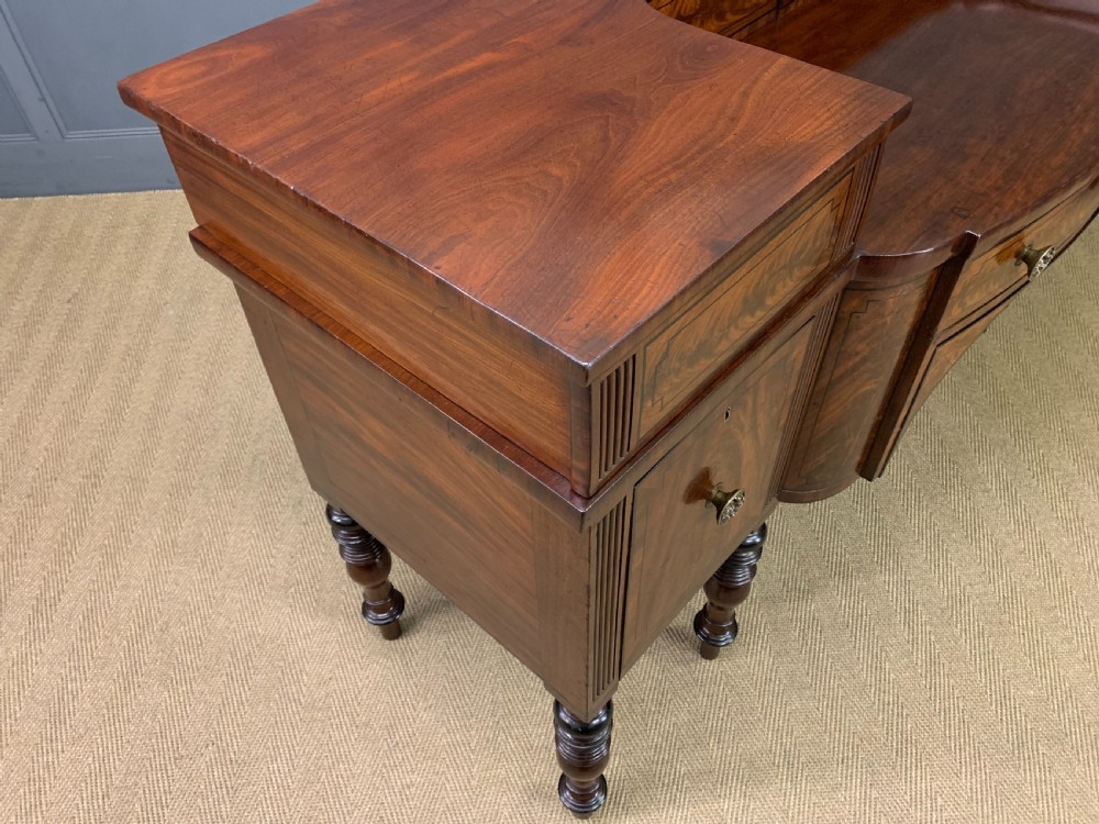 A Regency period flame mahogany sideboard featuring a raised stage top, central drawer, two side cupboards, detailed stringing, and turned reeded legs. View XXV
