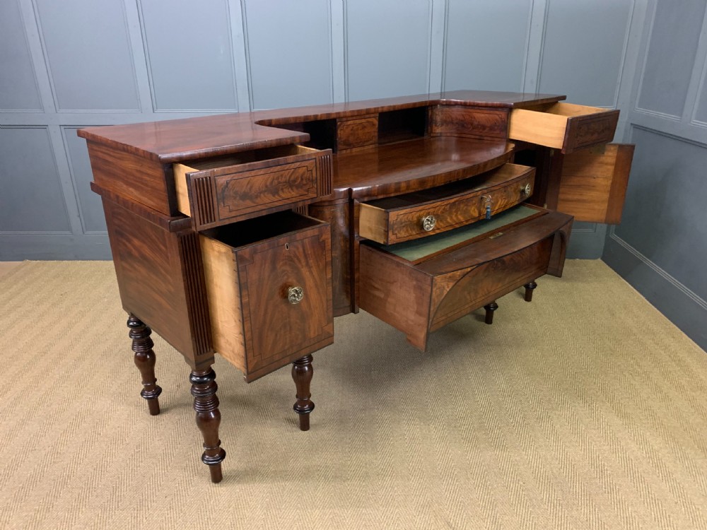 A Regency period flame mahogany sideboard featuring a raised stage top, central drawer, two side cupboards, detailed stringing, and turned reeded legs. View XXVI
