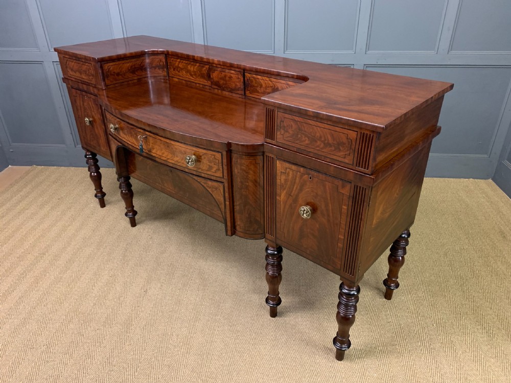 A Regency period flame mahogany sideboard featuring a raised stage top, central drawer, two side cupboards, detailed stringing, and turned reeded legs. View XXVII