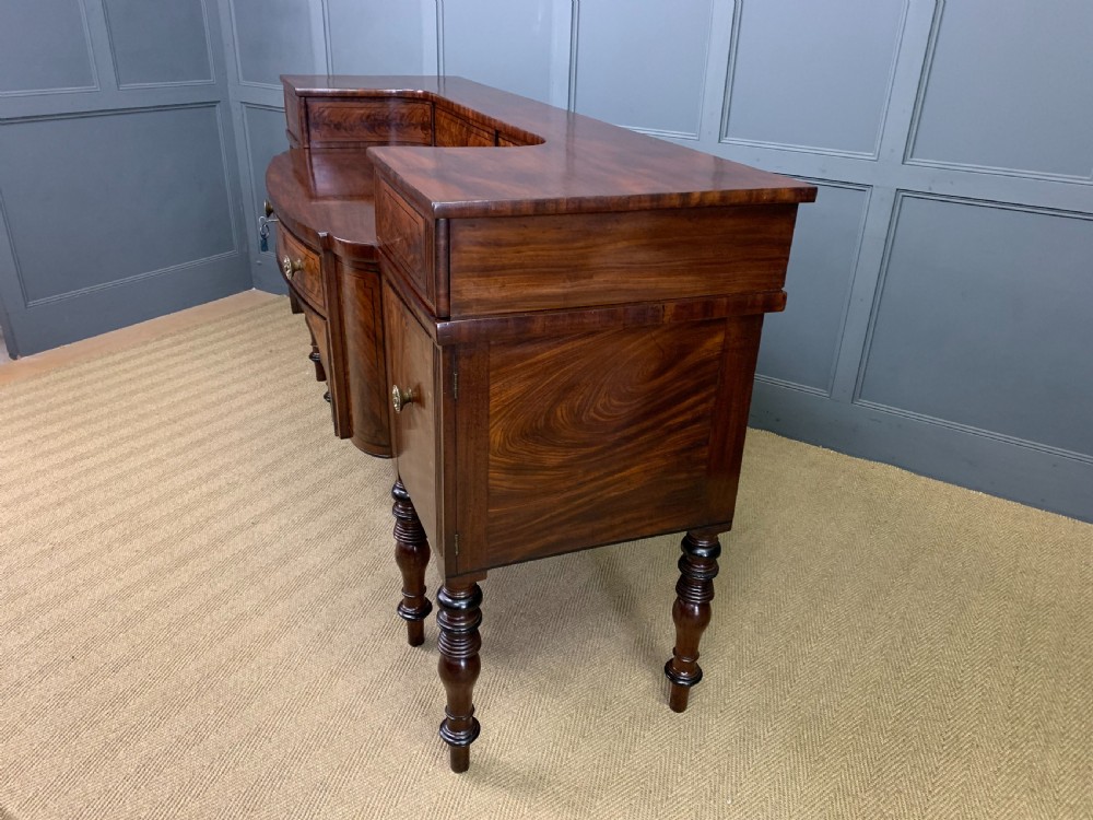 A Regency period flame mahogany sideboard featuring a raised stage top, central drawer, two side cupboards, detailed stringing, and turned reeded legs. View XXVIII