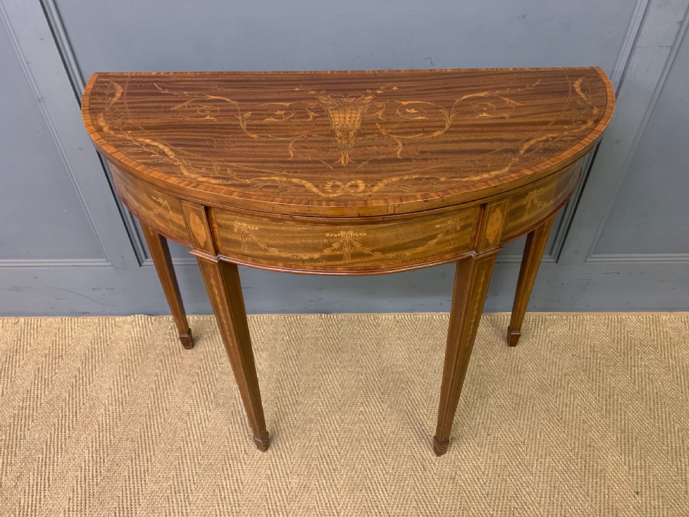 A demilune mahogany card table with floral marquetry inlays on the fold-over top and four square tapered legs.