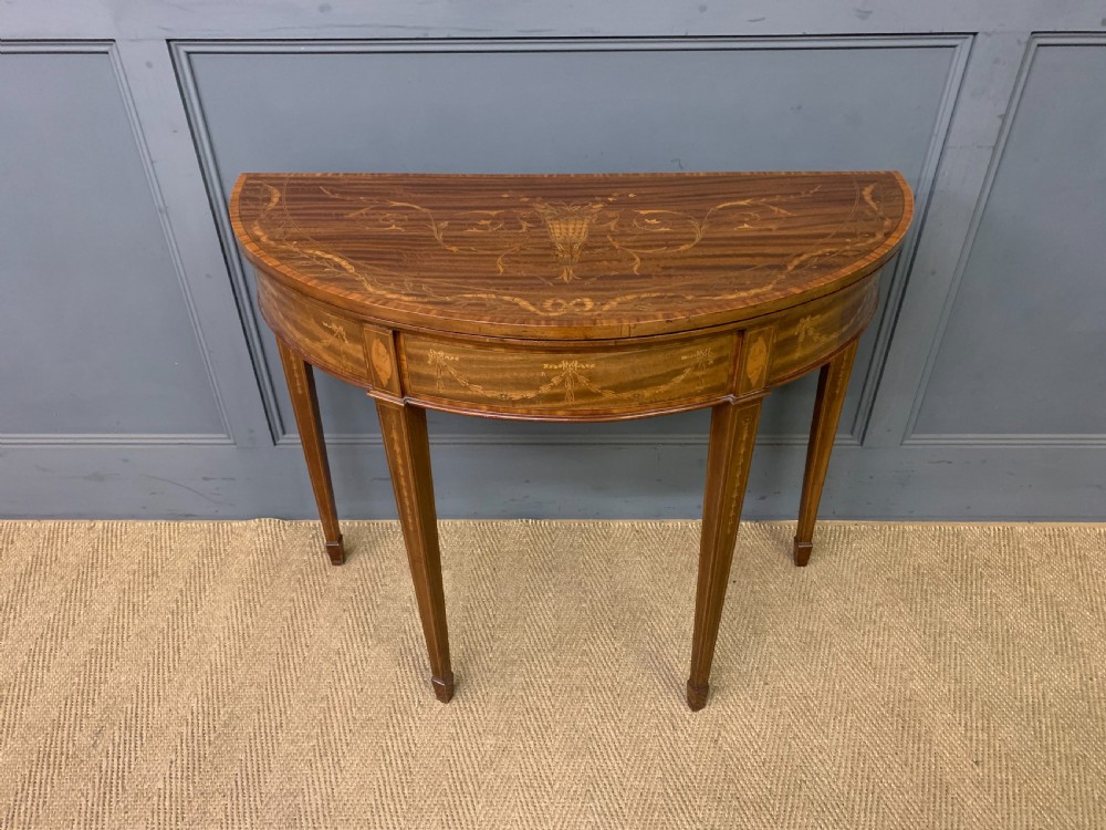 A demilune mahogany card table with floral marquetry inlays on the fold-over top and four square tapered legs. View II