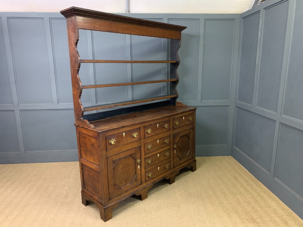 An early nineteenth-century Welsh oak dresser featuring a three-tier open plate rack above a base with drawers, cupboards, and brass hardware. View XV