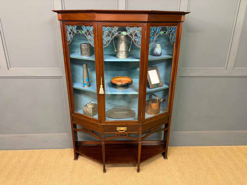 An Arts and Crafts mahogany display cabinet with stylized blue and green stained glass top panels, three glazed doors, and a blue fabric-lined interior.