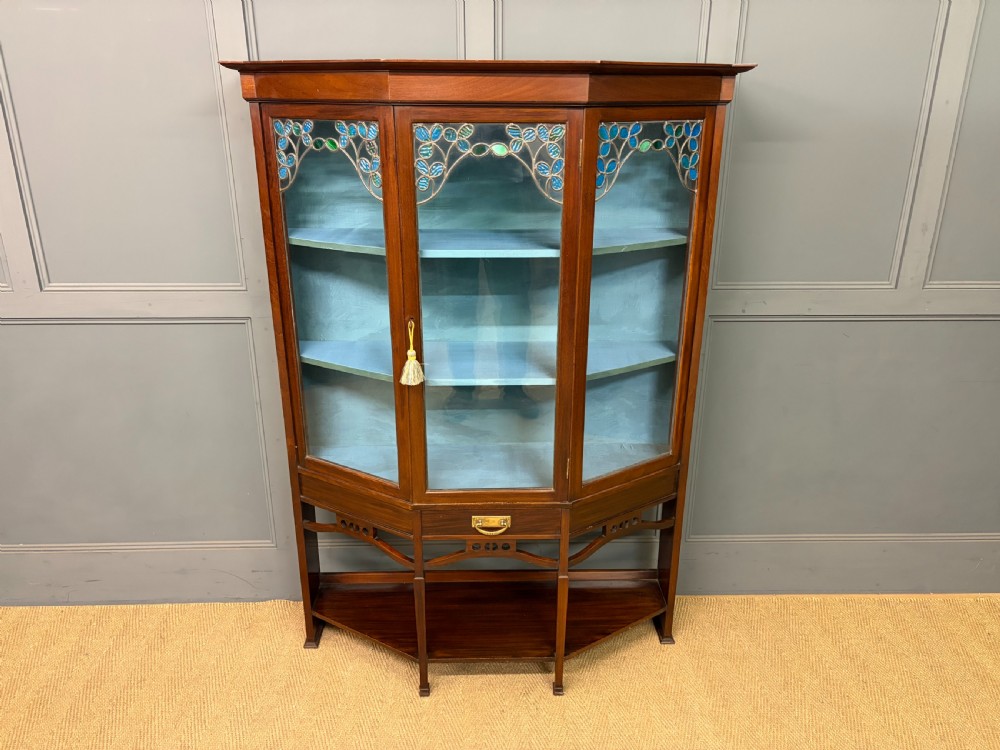An Arts and Crafts mahogany display cabinet with stylized blue and green stained glass top panels, three glazed doors, and a blue fabric-lined interior. View II