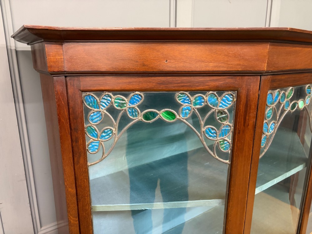 An Arts and Crafts mahogany display cabinet with stylized blue and green stained glass top panels, three glazed doors, and a blue fabric-lined interior. View VI
