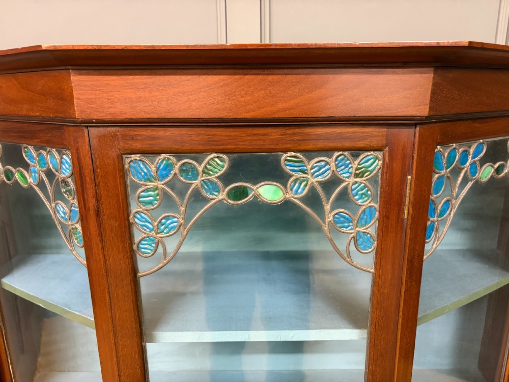 An Arts and Crafts mahogany display cabinet with stylized blue and green stained glass top panels, three glazed doors, and a blue fabric-lined interior. View VII