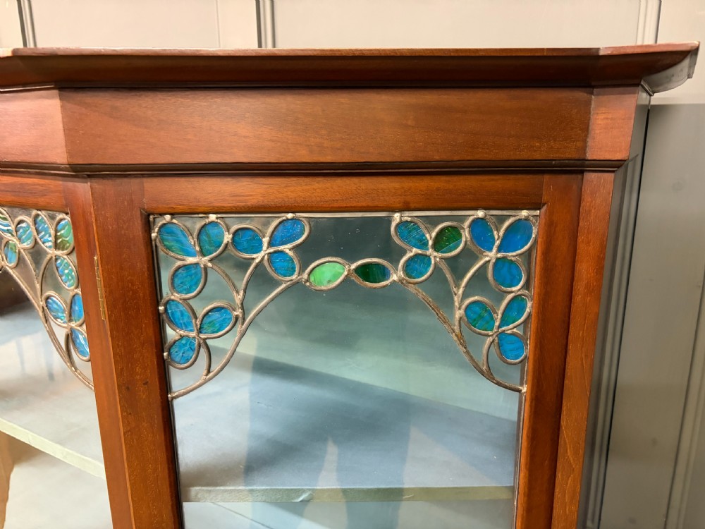 An Arts and Crafts mahogany display cabinet with stylized blue and green stained glass top panels, three glazed doors, and a blue fabric-lined interior. View VIII