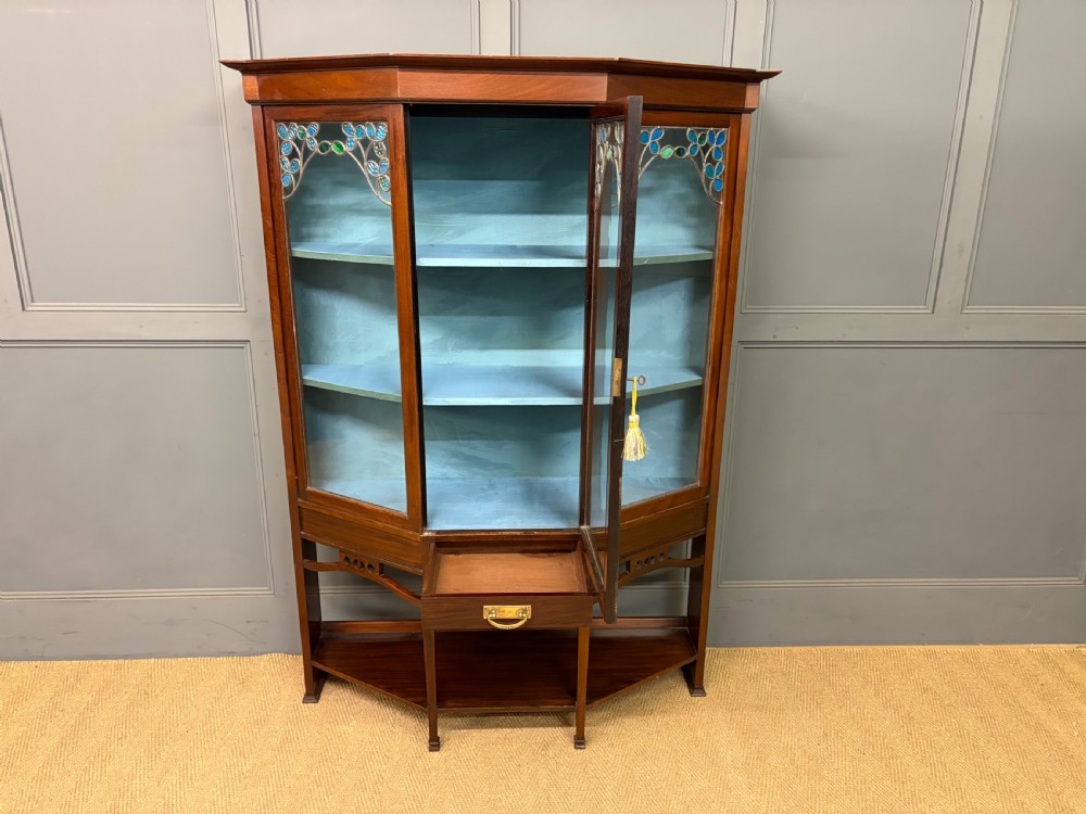 An Arts and Crafts mahogany display cabinet with stylized blue and green stained glass top panels, three glazed doors, and a blue fabric-lined interior. View IX