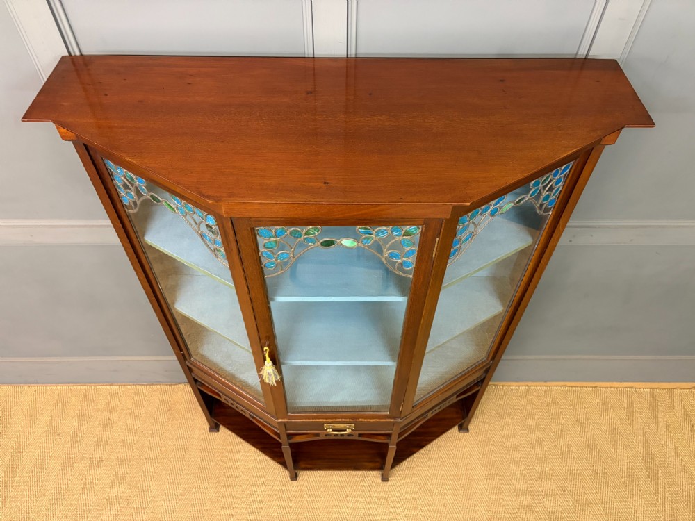 An Arts and Crafts mahogany display cabinet with stylized blue and green stained glass top panels, three glazed doors, and a blue fabric-lined interior. View X