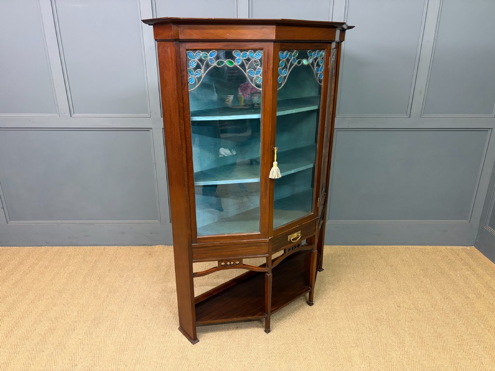 An Arts and Crafts mahogany display cabinet with stylized blue and green stained glass top panels, three glazed doors, and a blue fabric-lined interior. View XI