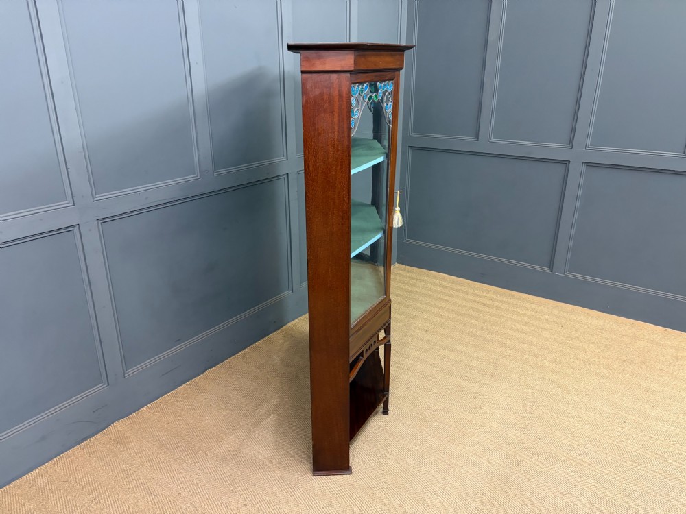 An Arts and Crafts mahogany display cabinet with stylized blue and green stained glass top panels, three glazed doors, and a blue fabric-lined interior. View XII