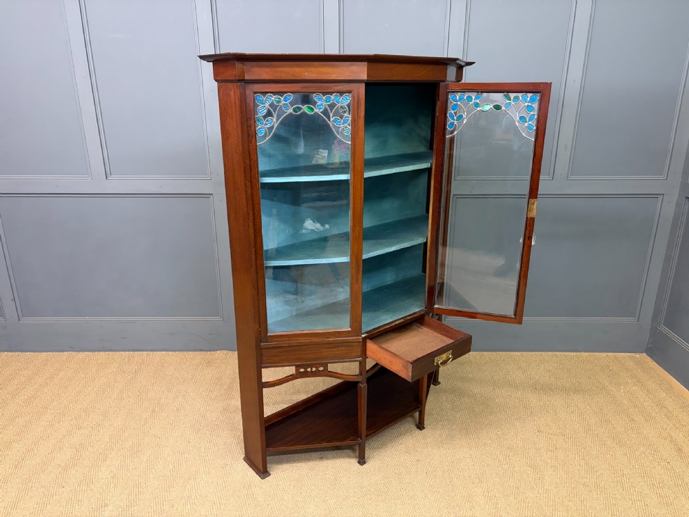 An Arts and Crafts mahogany display cabinet with stylized blue and green stained glass top panels, three glazed doors, and a blue fabric-lined interior. View XIII