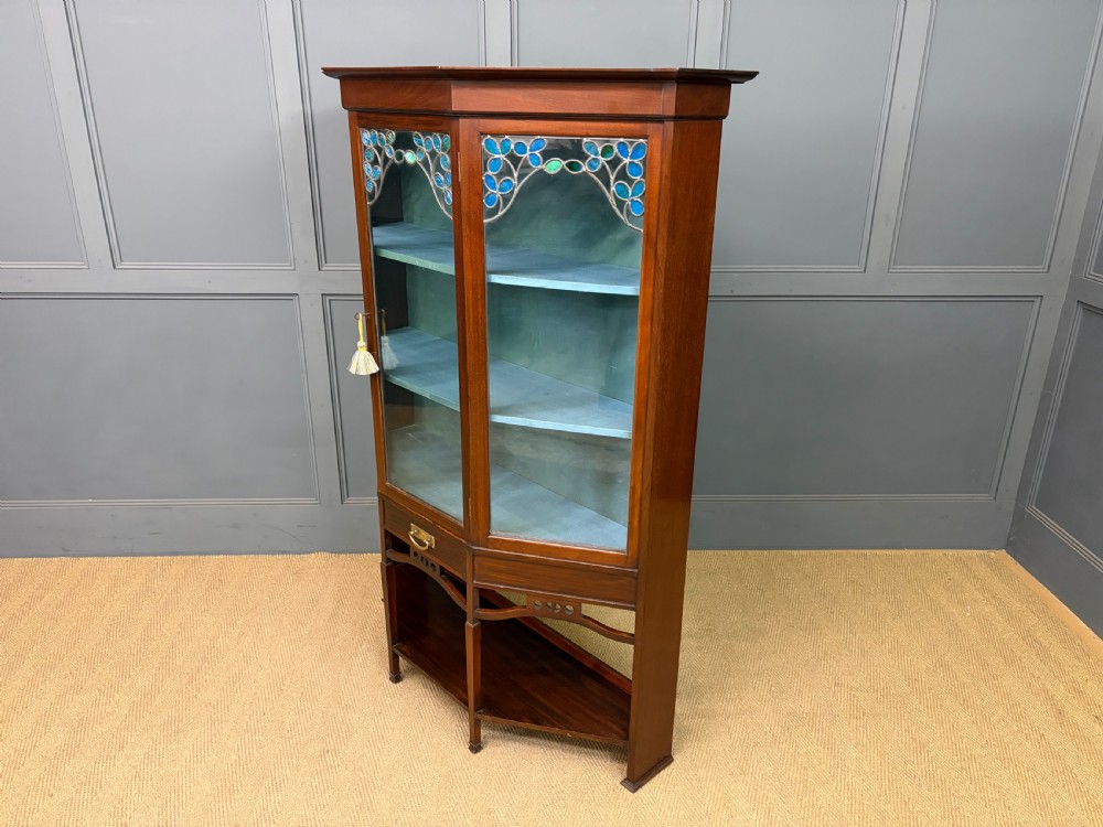 An Arts and Crafts mahogany display cabinet with stylized blue and green stained glass top panels, three glazed doors, and a blue fabric-lined interior. View XIV