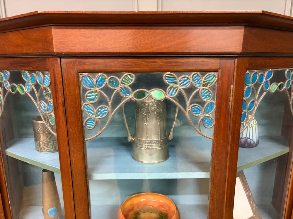An Arts and Crafts mahogany display cabinet with stylized blue and green stained glass top panels, three glazed doors, and a blue fabric-lined interior. View XV