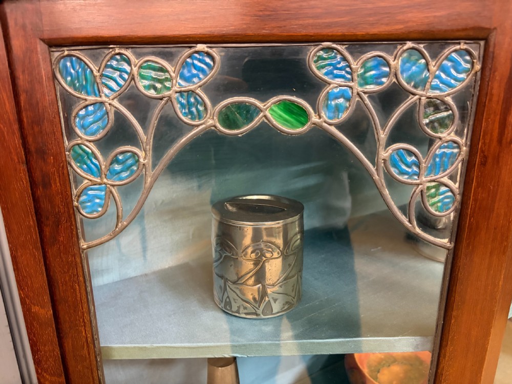 An Arts and Crafts mahogany display cabinet with stylized blue and green stained glass top panels, three glazed doors, and a blue fabric-lined interior. View XVI