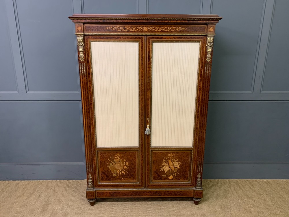A 19th-century French double-door bookcase featuring amboyna and mahogany veneers, intricate floral marquetry, pleated cream fabric door panels, and brass mounts.