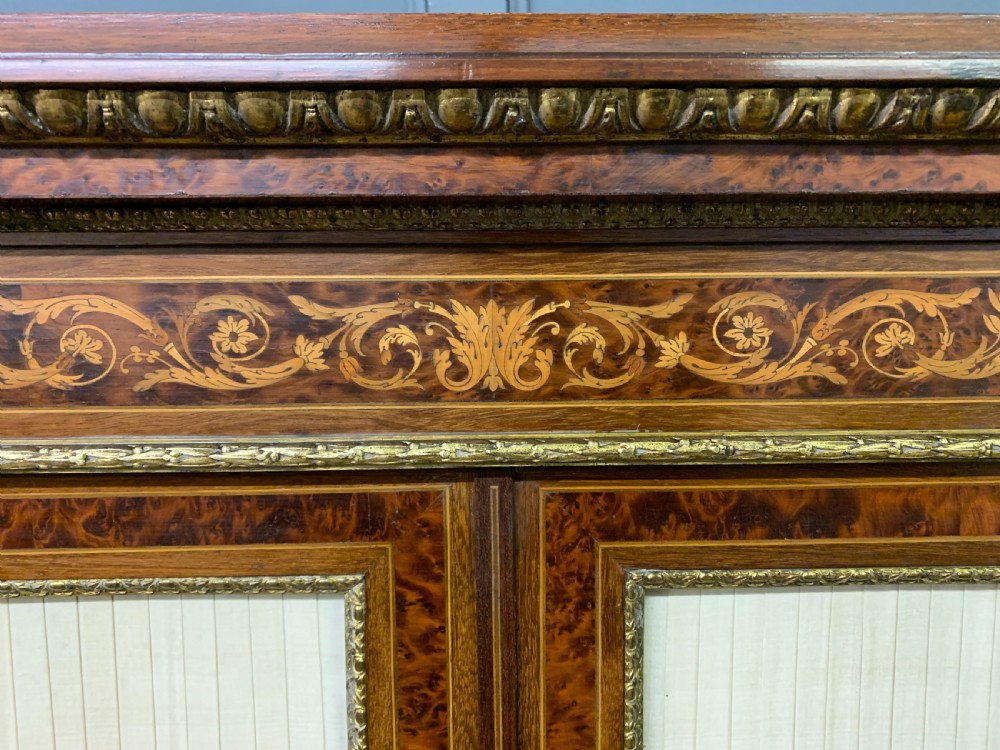 A 19th-century French double-door bookcase featuring amboyna and mahogany veneers, intricate floral marquetry, pleated cream fabric door panels, and brass mounts. View XIX
