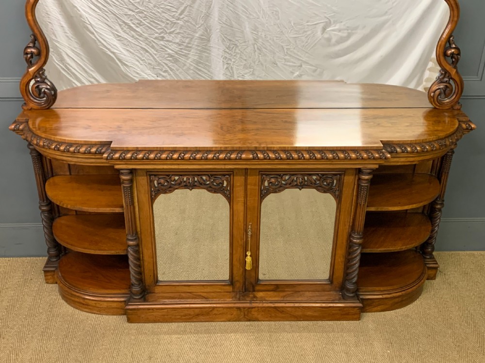 A Victorian burr walnut chiffonier featuring open side display shelves, mirrored central cabinet doors, and an ornate carved mirror frame cresting. View V