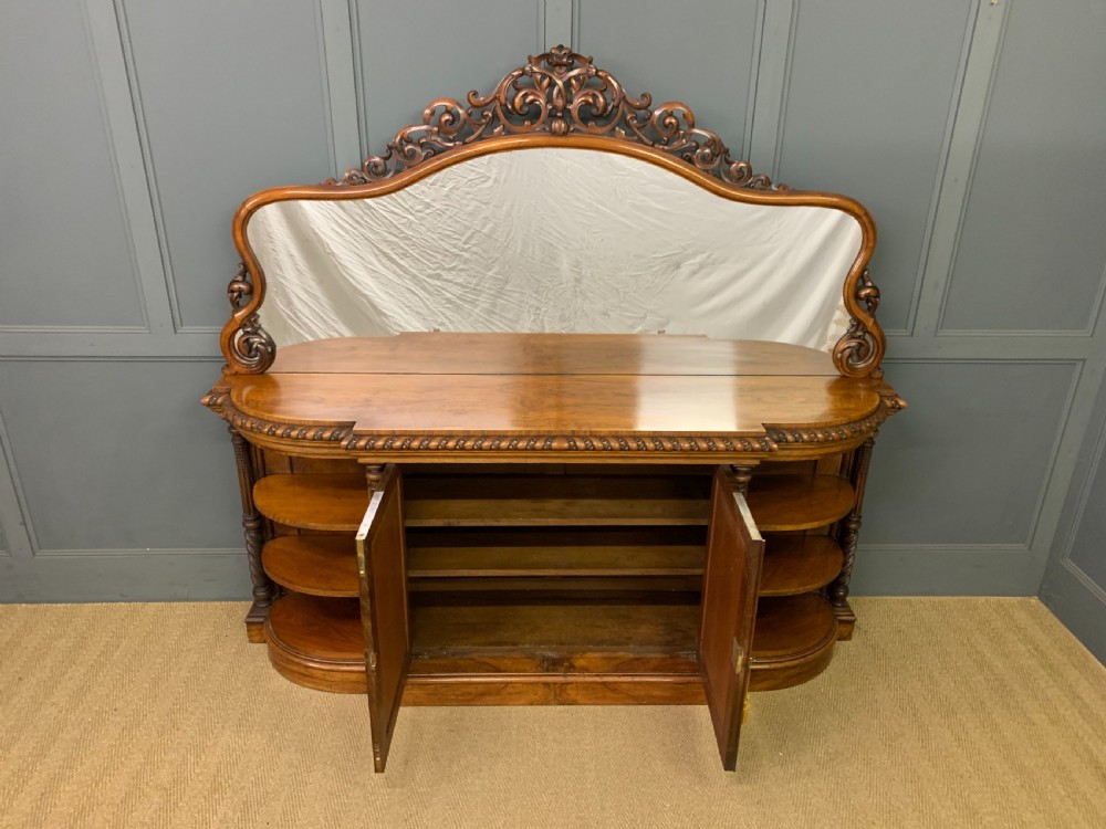 A Victorian burr walnut chiffonier featuring open side display shelves, mirrored central cabinet doors, and an ornate carved mirror frame cresting. View VI