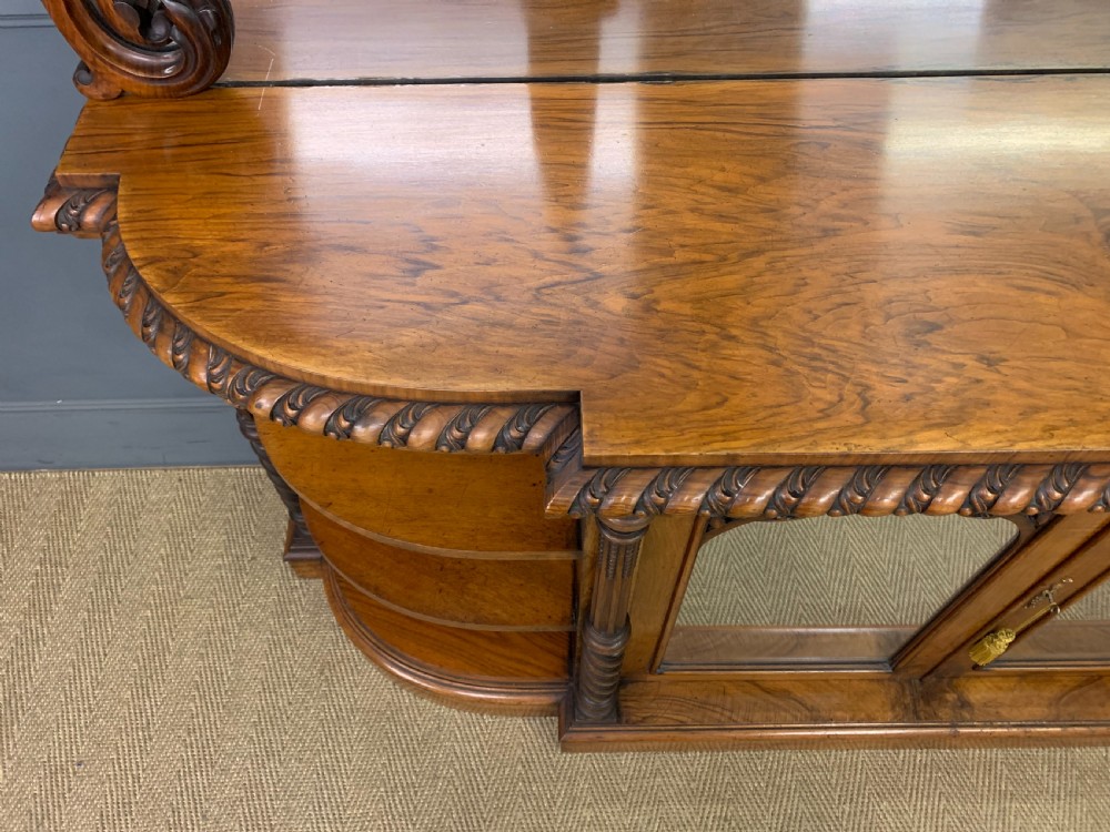 A Victorian burr walnut chiffonier featuring open side display shelves, mirrored central cabinet doors, and an ornate carved mirror frame cresting. View XV
