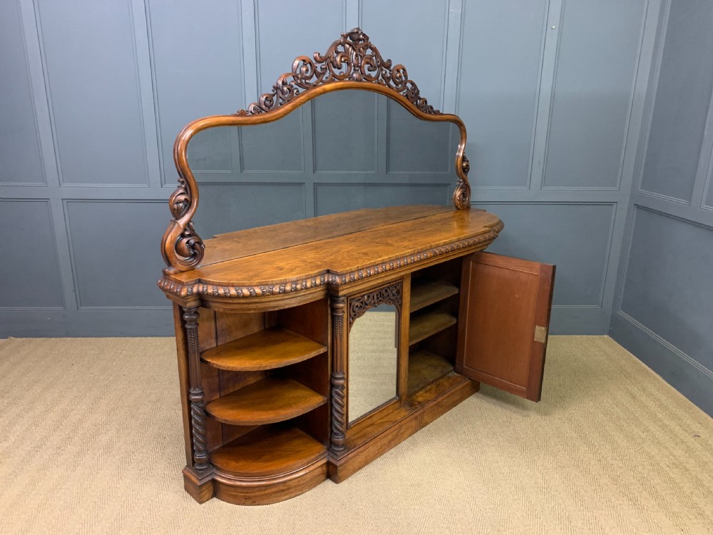A Victorian burr walnut chiffonier featuring open side display shelves, mirrored central cabinet doors, and an ornate carved mirror frame cresting. View XIX