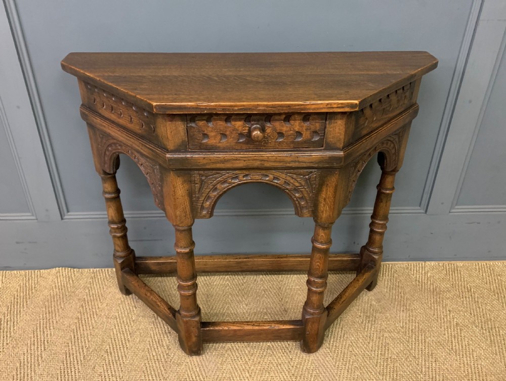 A carved dark oak side table featuring a canted five-sided top, relief carved arches on the frieze, a single drawer, turned legs, and a lower stretcher base.