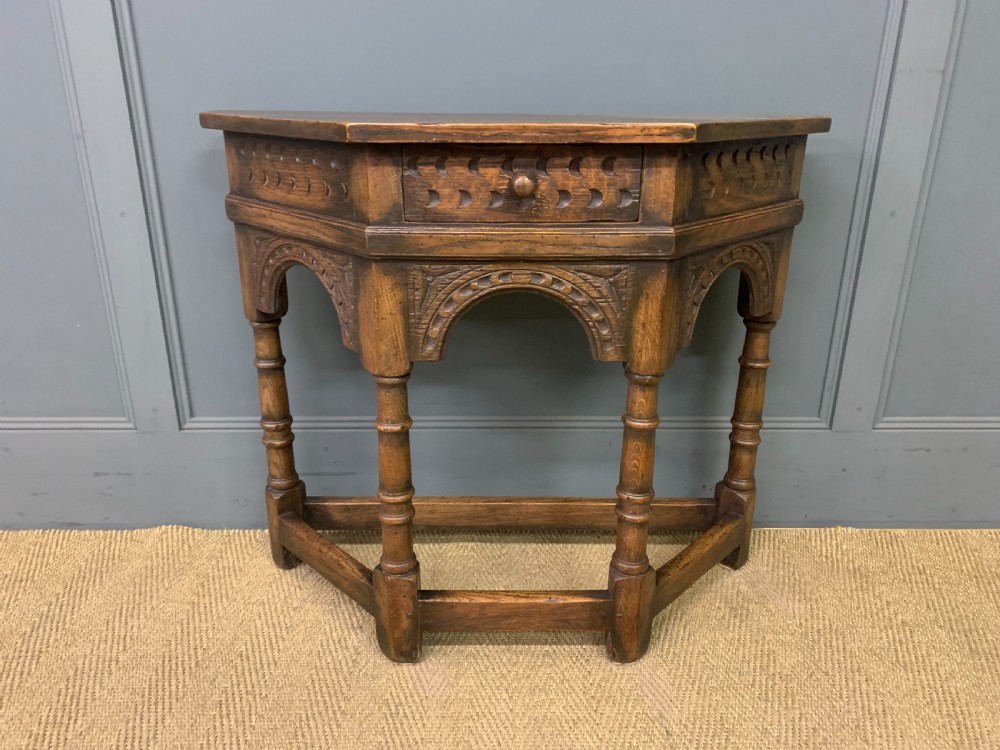 A carved dark oak side table featuring a canted five-sided top, relief carved arches on the frieze, a single drawer, turned legs, and a lower stretcher base. View II