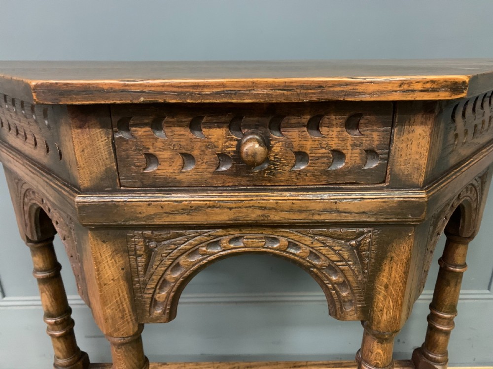 A carved dark oak side table featuring a canted five-sided top, relief carved arches on the frieze, a single drawer, turned legs, and a lower stretcher base. View IV