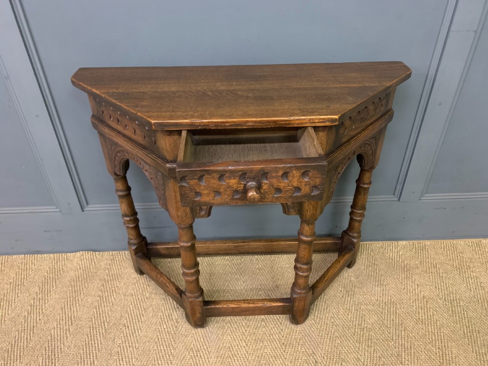 A carved dark oak side table featuring a canted five-sided top, relief carved arches on the frieze, a single drawer, turned legs, and a lower stretcher base. View V