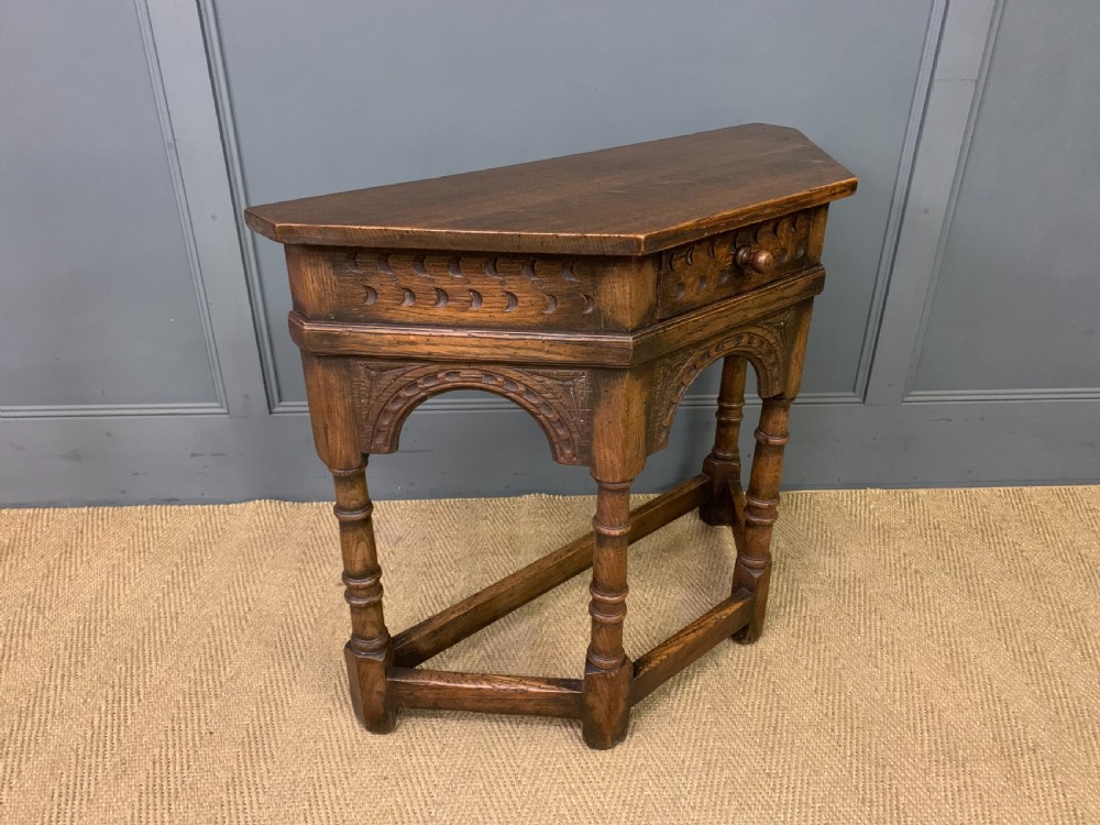 A carved dark oak side table featuring a canted five-sided top, relief carved arches on the frieze, a single drawer, turned legs, and a lower stretcher base. View VII