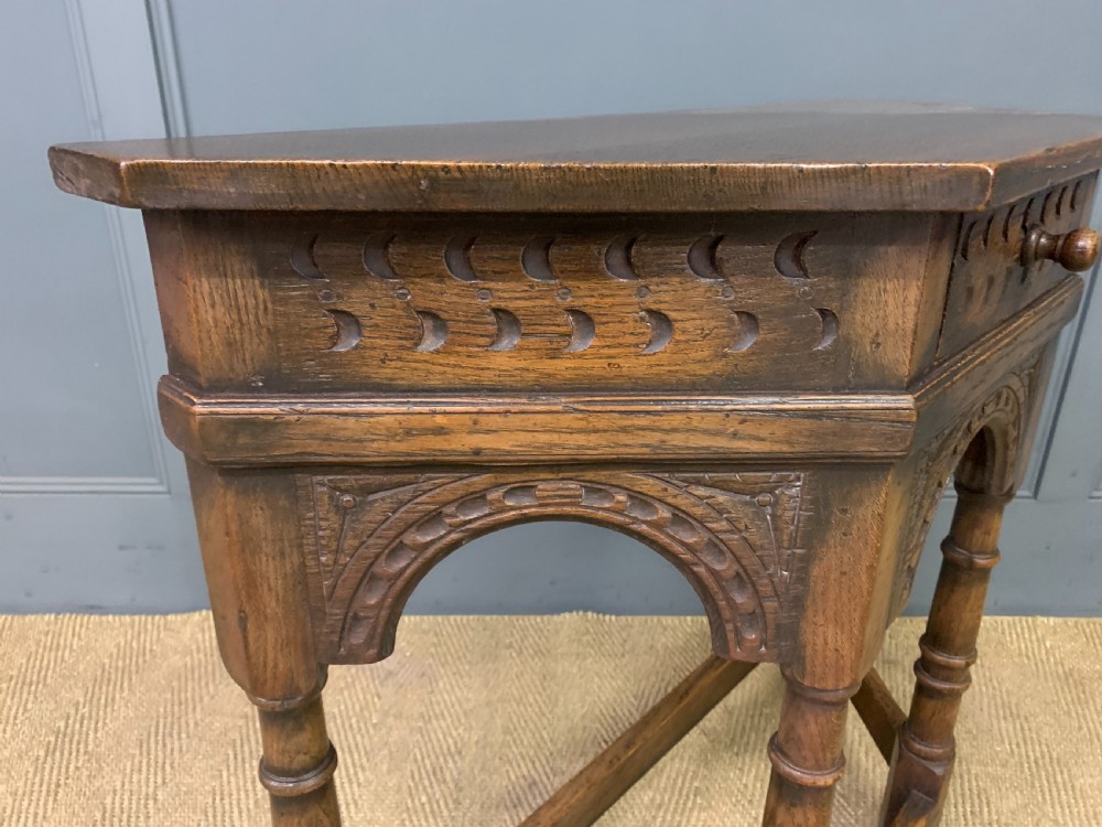 A carved dark oak side table featuring a canted five-sided top, relief carved arches on the frieze, a single drawer, turned legs, and a lower stretcher base. View X