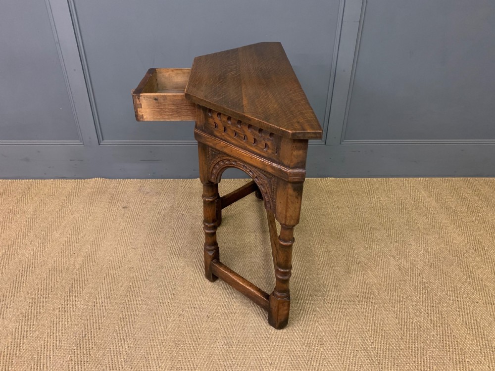 A carved dark oak side table featuring a canted five-sided top, relief carved arches on the frieze, a single drawer, turned legs, and a lower stretcher base. View XII