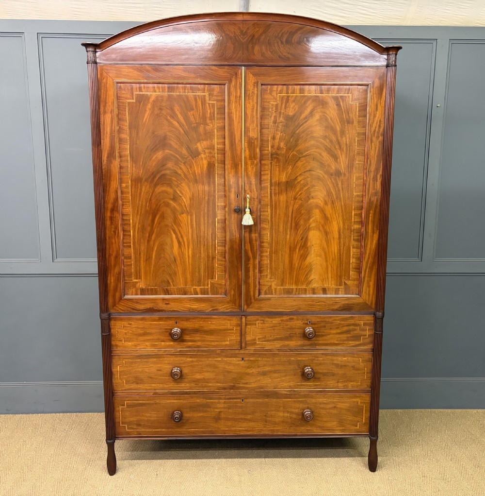 An antique Georgian flame mahogany linen press with an arched top cornice, two inlaid cupboard doors, fluted side pilasters, and a base of five drawers with turned knobs.